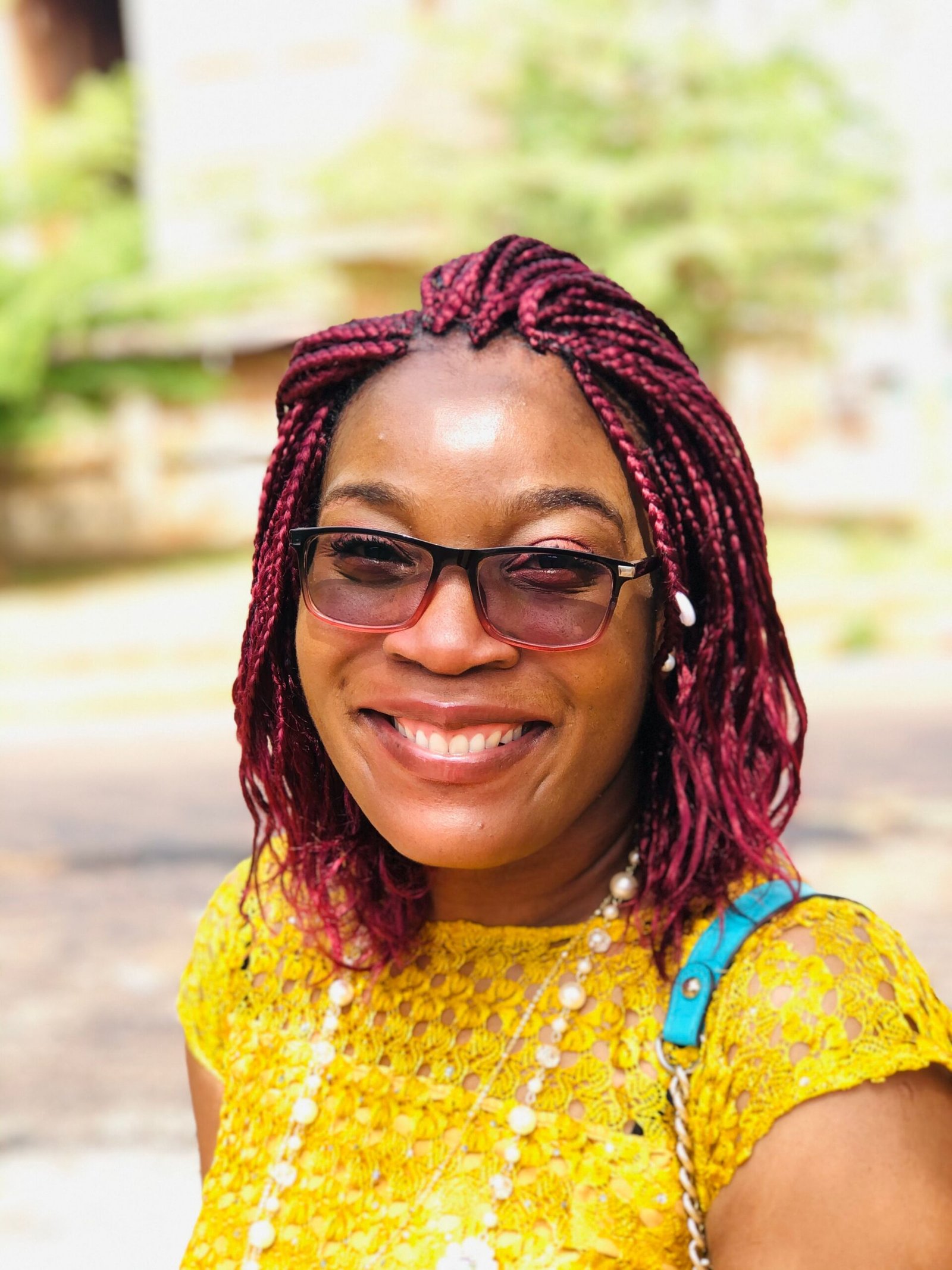 A young woman smiles widely at the camera. She is outside, wearing bright summer clothes and accessories.