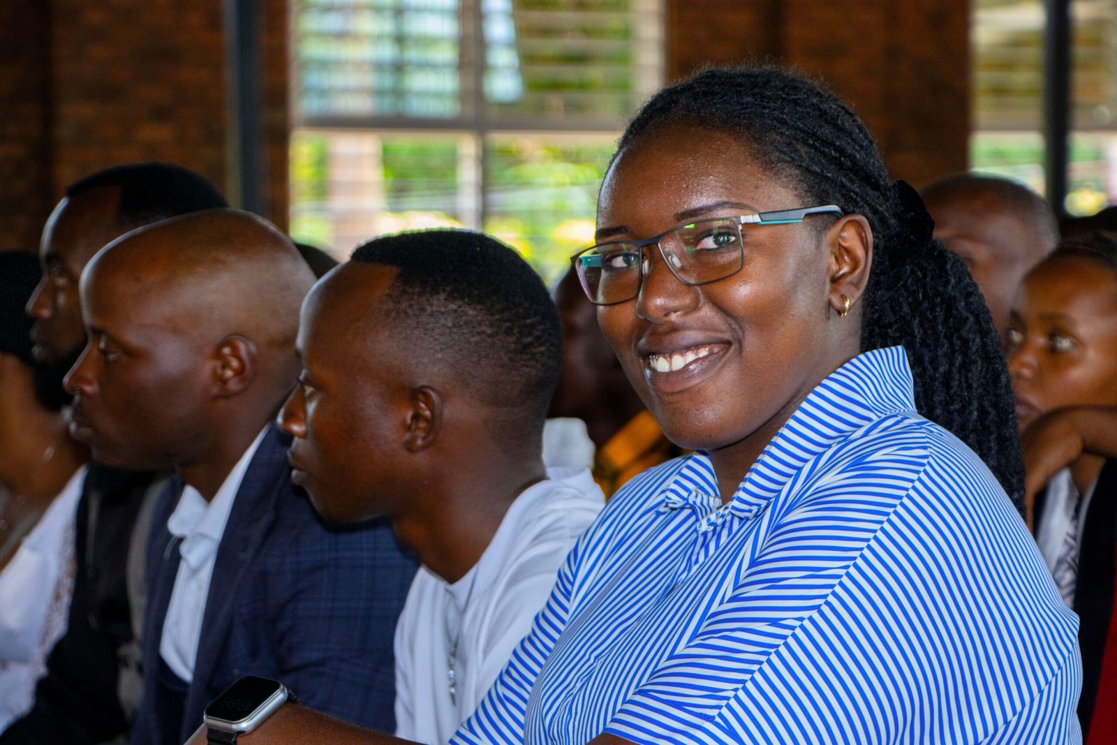 A young woman smiles at the camera. Around her, many young men look towards the left, at what seems to be the front of a classroom.