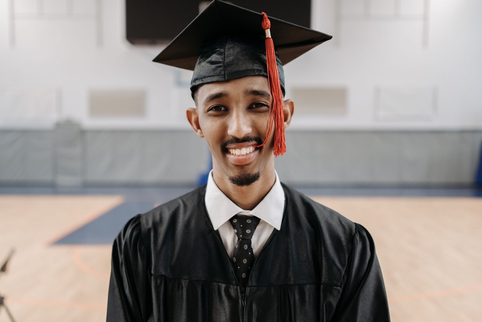 A young man smiles at the camera wearing formal graduation attire. In the background there is a gym.