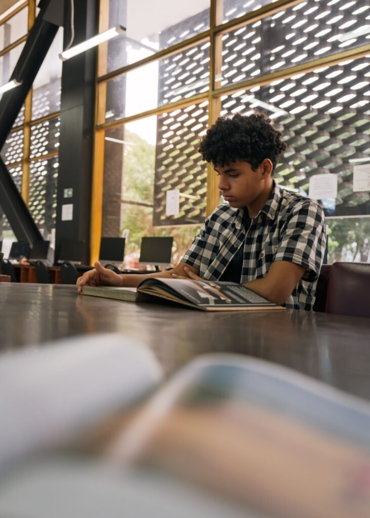 A young man in casual clothes sits on a big table reading a book. Within the frame, there is another book in front of him and a big window behind him.